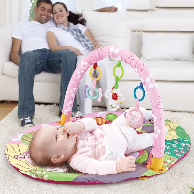 תינוקת על משטח פעילות עגול עם קשת ורודה ותליונים, הורים ברקע על ספה